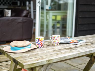 A wooden picnic table with two colorful mugs a sun hat and magazines on a patio outside a house