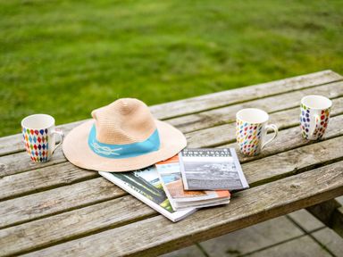 A wooden picnic table outside with three patterned mugs a straw hat and guidebooks on it at Dolphin Lodge in Port Isaac