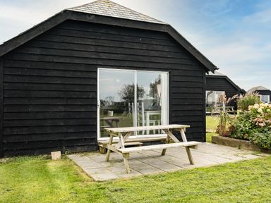 A wooden picnic table on a patio outside a black wooden building with sliding glass doors at Dolphin Lodge in Port Isaac