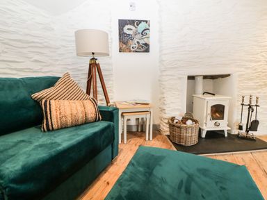 A room with a green sofa a floor lamp a wood stove with tools and basket tables and artwork on a white textured wall at Spindrift in Polperro