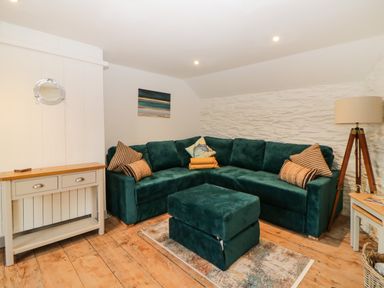 A living room with a green sectional sofa a matching ottoman a wooden floor a side table a tripod floor lamp and a small console table at Spindrift in Polperro