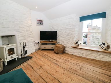 A corner of a room with a television on a wooden stand a wood stove with fireplace tools a window with a shelf and cushions and wooden floor at Spindrift in Polperro