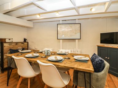 A dining room with a wooden table set for six with white and grey chairs a map on the wall and a television on a cabinet at Spindrift in Polperro