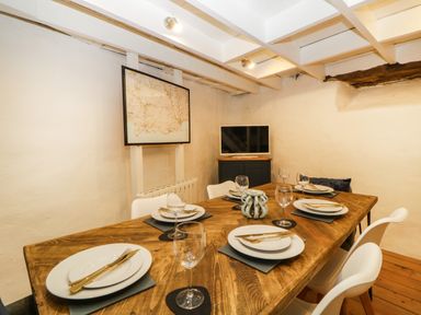 A dining room with a wooden table set with plates glasses and cutlery white chairs TV and a framed map on the wall at Spindrift in Polperro