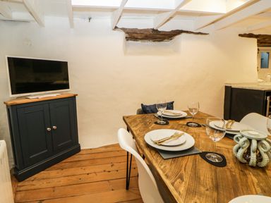 A dining area with a wooden table set with plates wine glasses and cutlery a small cabinet with a television on top in a room with exposed wooden beams at Spindrift in Polperro