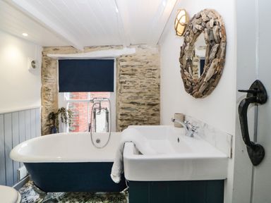 A bathroom with a freestanding bathtub a sink a round wooden mirror and a window with a dark blind at Spindrift in Polperro