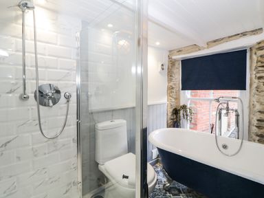 A bathroom with a shower, toilet, freestanding bathtub, window with a dark blind, and exposed stone walls at Spindrift in Polperro