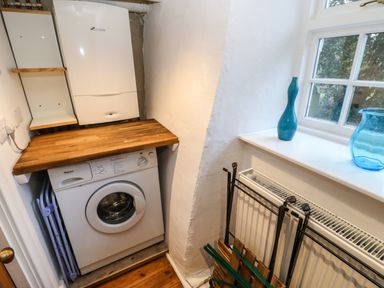 A laundry area with a washing machine under a wooden countertop and a boiler on the wall next to a window with decorative vases at Cross View Cottage in Alston