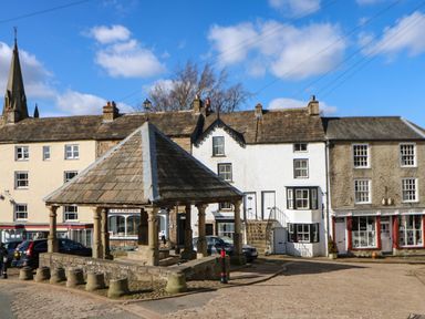 An outdoor market shelter with a pitched roof surrounded by stone bollards in a town square with historic stone buildings and cars parked nearby at Cross View Cottage in Alston