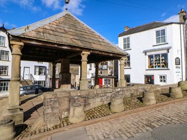 A stone market cross with pillars and a tiled roof on a cobblestone street at Cross View Cottage in Alston
