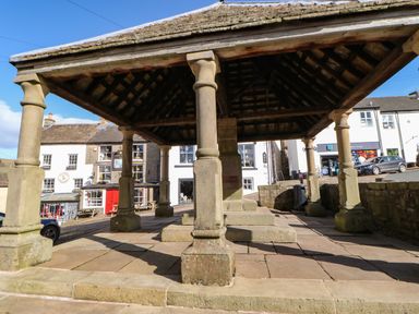 A stone pavilion with columns and a pitched roof in a town square with buildings and cars in the background at Cross View Cottage in Alston