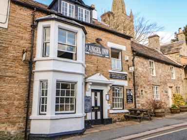 Exterior view of a stone building with signs for The Angel Inn and a wooden bench outside at Cross View Cottage in Alston