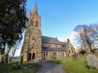 A stone church with a tall steeple surrounded by gravestones and trees at Cross View Cottage in Alston