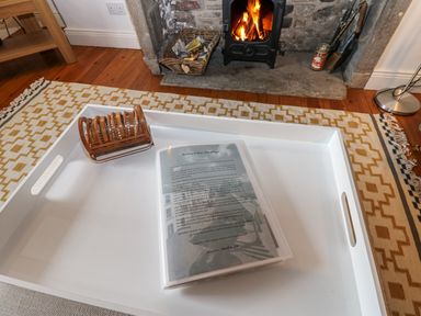 A white tray on a patterned rug with a booklet and a wooden napkin holder in front of a fireplace with a burning fire at Cross View Cottage in Alston