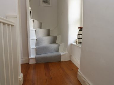 A narrow staircase with a grey carpet runner and a wooden floor next to a small window with a ceramic jar at Cross View Cottage in Alston