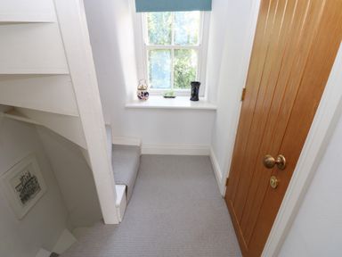 A small carpeted landing with a wooden door white stairs and a window with decorative items on the sill at Cross View Cottage in Alston
