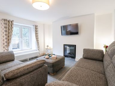 A living room with grey sofas a window with curtains a wall-mounted TV and an electric fireplace at Beach Road House in Hele Bay Ilfracombe