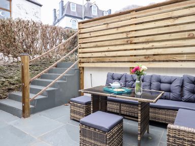 An outdoor patio area with rattan furniture including a table with a tea set and flowers a wooden slatted fence and stairs with rope handrails at Beach Road House Hele Bay Ilfracombe