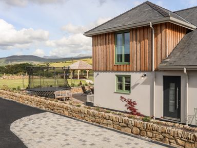 An exterior view of a house with a wooden upper facade a paved pathway a trampoline and a gazebo in the garden at Rynys in Llanddeiniolen near Llanrug