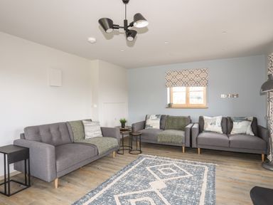 A living room with three grey sofas a patterned rug and a window with a shade at Rynys in Llanddeiniolen near Llanrug
