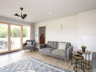 A living room with a grey sofa and armchair a patterned rug wooden nesting tables and glass doors at Rynys in Llanddeiniolen near Llanrug