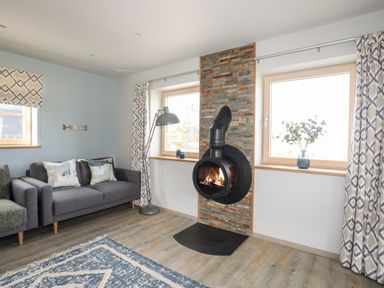 A living room with a gray sofa a hanging black fireplace and two windows with curtains at Rynys in Llanddeiniolen near Llanrug