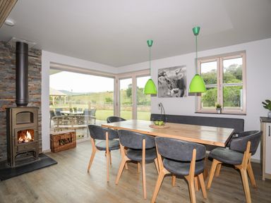 A dining room with a wooden table and chairs a wood burning stove and large windows showing an outdoor patio and fields at Rynys in Llanddeiniolen near Llanrug