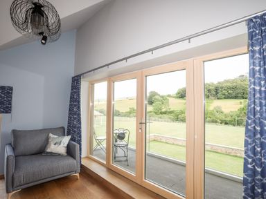 A sitting area with a gray armchair and large glass doors opening to a patio and green fields at Rynys in Llanddeiniolen near Llanrug