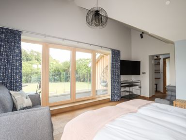 A bedroom with a bed a gray chair a television on a stand and large windows with blue curtains at Rynys in Llanddeiniolen near Llanrug