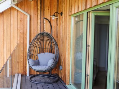 An outdoor wooden balcony with a hanging egg chair with cushions and a green-framed glass door at Rynys in Llanddeiniolen near Llanrug