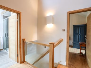 A hallway with wooden floors a staircase with glass and wood railing two open doors one leading to a bathroom and the other to a room with a blue wardrobe and wooden table at Rynys in Llanddeiniolen near Llanrug