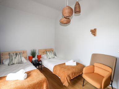A bedroom with two single beds with orange blankets and pillows a small table with lamps and a plant and an orange armchair at Rynys in Llanddeiniolen near Llanrug