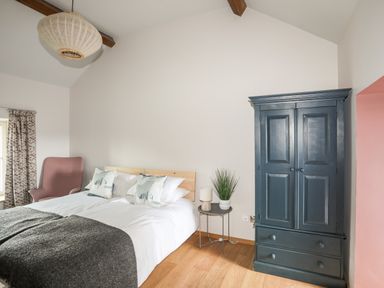 A bedroom with a bed pillows a side table plant lamp a blue wardrobe and a pink chair at Rynys in Llanddeiniolen near Llanrug