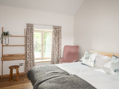 A bedroom with a bed pillows a pink armchair a wooden stool a window with patterned curtains and a wooden desk at Rynys in Llanddeiniolen near Llanrug