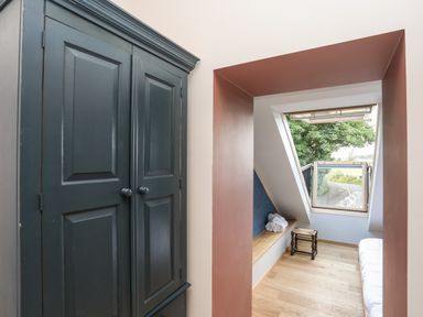 A room with a dark wardrobe wooden stool bench with towels and an open window overlooking a road at Rynys in Llanddeiniolen near Llanrug