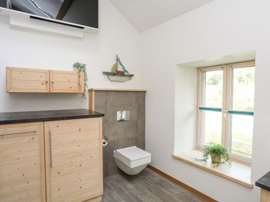 A bathroom with a wall-mounted toilet wooden cabinets a window with a plant and a decorative sailboat on the wall at Rynys in Llanddeiniolen near Llanrug