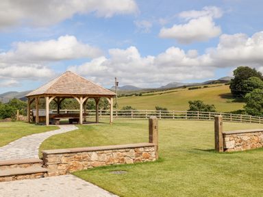 A wooden gazebo with benches on a lawn with stone steps and a fence in a countryside setting at Rynys in Llanddeiniolen near Llanrug