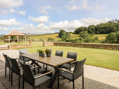 An outdoor patio with a dining table and chairs overlooking a lawn and hills at Rynys in Llanddeiniolen near Llanrug