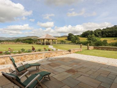 A patio with wooden chairs and stone flooring overlooking a garden with a gazebo and hills at Rynys in Llanddeiniolen near Llanrug
