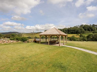 A wooden gazebo with a picnic table on a lawn with hills and trees in the background at Rynys in Llanddeiniolen near Llanrug