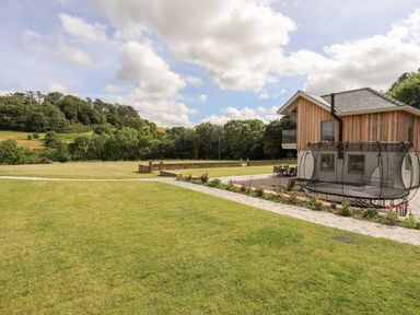 A grass lawn with a stone path leading to a modern wooden house with a trampoline and outdoor seating at Rynys in Llanddeiniolen near Llanrug