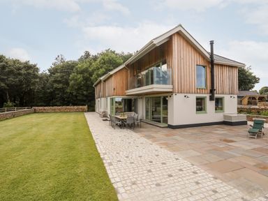 A modern two-story house with wood paneling and large windows next to a stone patio and lawn at Rynys in Llanddeiniolen near Llanrug
