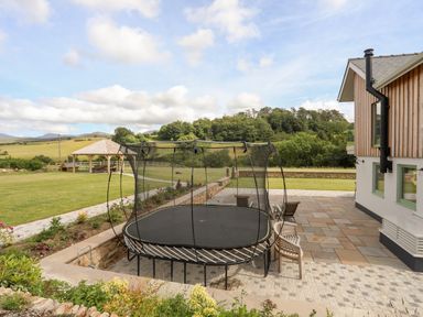 An outdoor patio with a trampoline surrounded by chairs and a garden with a gazebo and trees in the background at Rynys in Llanddeiniolen near Llanrug