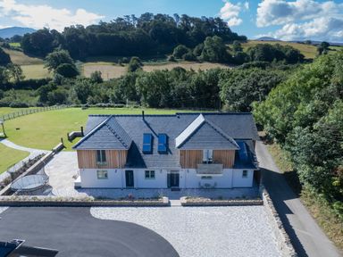 A modern detached house with wood and white exterior walls and a slate roof surrounded by trees and fields at Rynys in Llanddeiniolen near Llanrug