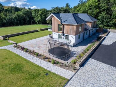 A two storey house with a trampoline on the patio and a garden surrounded by trees at Rynys in Llanddeiniolen near Llanrug