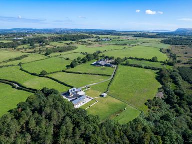An aerial view of green fields with a house and farm buildings surrounded by trees at Rynys in Llanddeiniolen near Llanrug