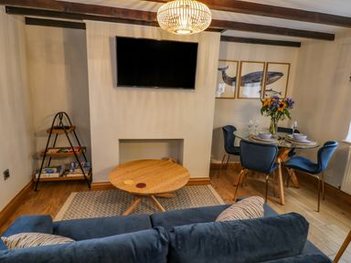 A living room with a sofa a round wooden coffee table a glass dining table with blue chairs and a wall mounted TV at April Cottage in Whitby