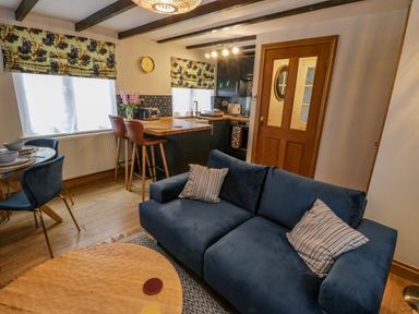 A living area with a blue sofa, a dining table with chairs, a kitchen island with bar stools, windows with patterned blinds, and a wooden door at April Cottage in Whitby
