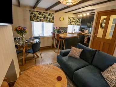 A living area with a blue sofa, round wooden coffee table, dining table with chairs and flowers, kitchen with bar stools and wooden door at April Cottage in Whitby