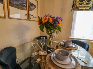 A dining table set with plates bowls glasses and gold cutlery with a flower vase and blue chairs at April Cottage in Whitby